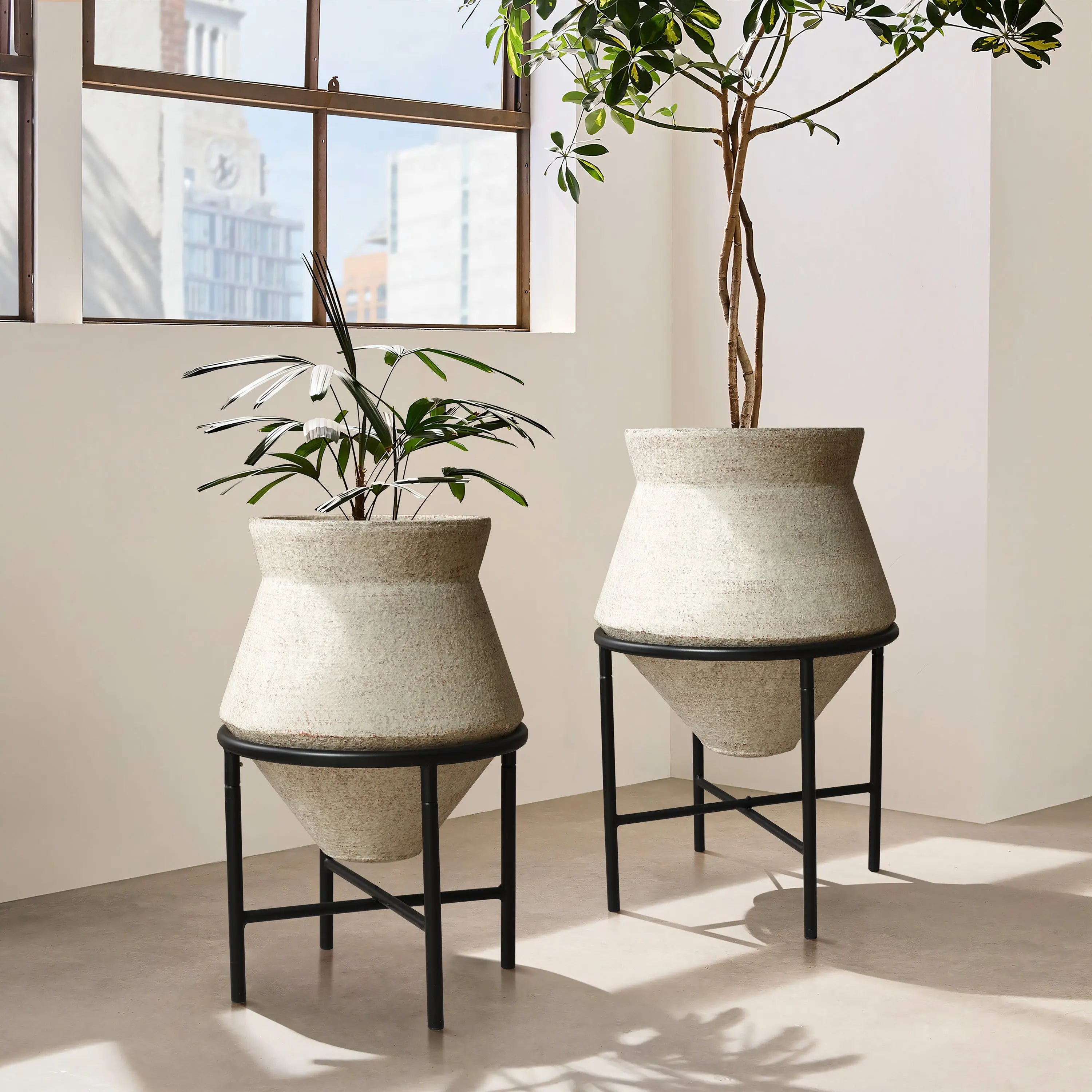Two large, beige cachepot planters on black metal stands hold tall, leafy plants, adding a touch of vintage distressed style to the sunlit room. They are beautifully placed in front of large windows, enhancing the indoor space decor with their textured charm.