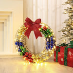 The holiday wreath, adorned with colorful baubles and twinkling LED lights, boasts a red bow. Nestled near the Christmas tree and surrounded by wrapped gifts on the floor, it creates a charming outdoor holiday decoration.
