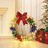 The holiday wreath, adorned with colorful baubles and twinkling LED lights, boasts a red bow. Nestled near the Christmas tree and surrounded by wrapped gifts on the floor, it creates a charming outdoor holiday decoration.