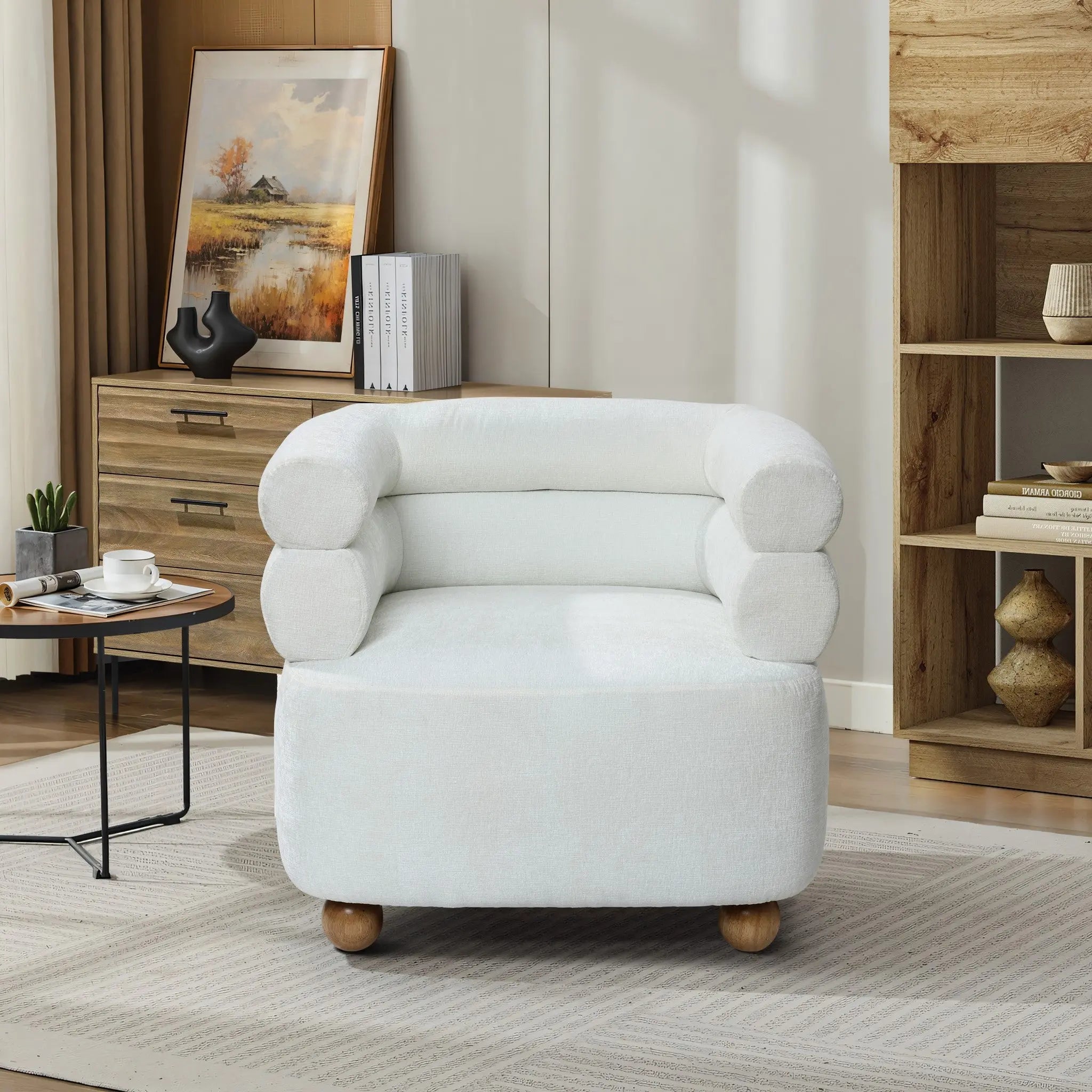 A cozy living room showcases the LuxenHome Tia ivory barrel accent chair with a double bolster backrest and rounded armrests on wooden ball legs, accompanied by a wooden cabinet, decorative vase, painting, books, and a round coffee table on a patterned rug.