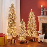 Three pre-lit artificial trees with lifelike PVC branches and gold ornaments stand beside a fireplace, surrounded by gift boxes and a decorative reindeer, creating a winter wonderland scene on a wooden floor against a red wall.