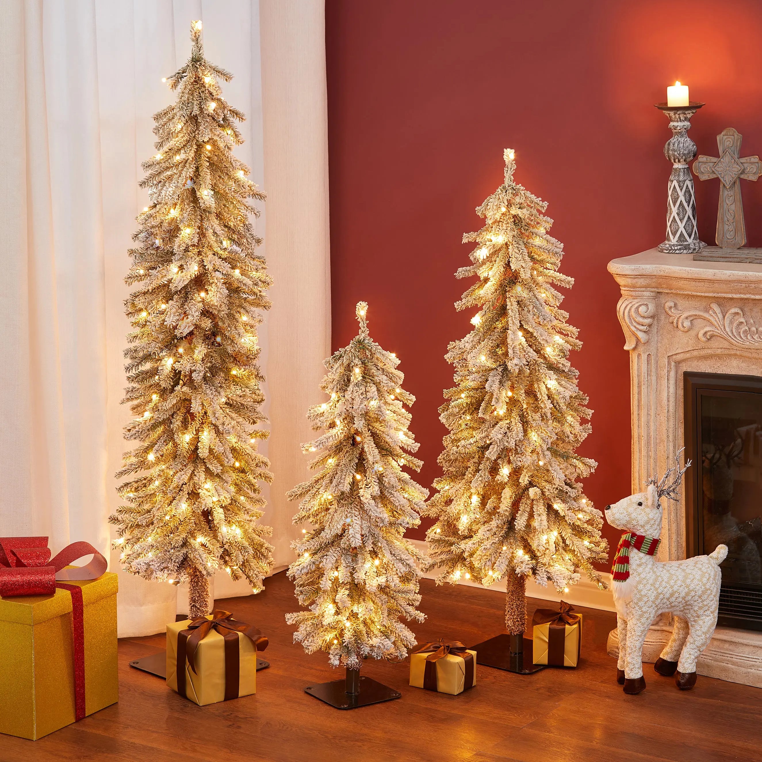 Three pre-lit artificial trees with lifelike PVC branches and gold ornaments stand beside a fireplace, surrounded by gift boxes and a decorative reindeer, creating a winter wonderland scene on a wooden floor against a red wall.