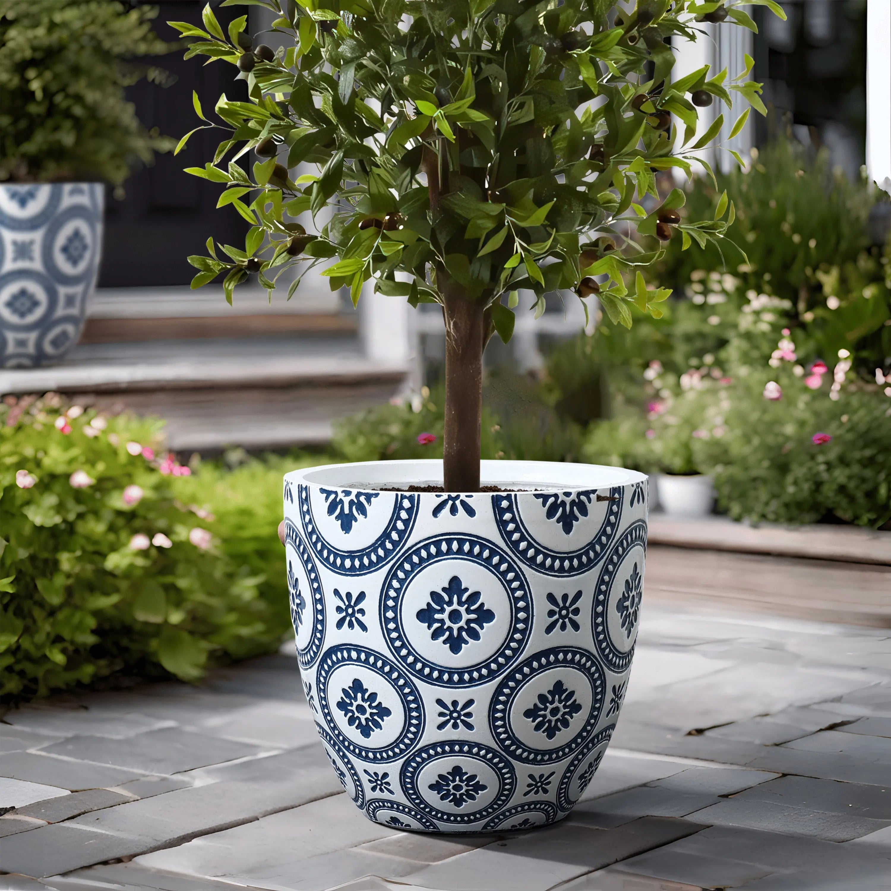 The decorative blue and white patterned pot, made of MgO material, cradles a small tree on the paved patio, surrounded by vibrant greenery and flowers.