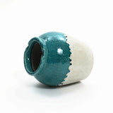 A ceramic vase with a glossy teal upper half and a textured beige lower half, embodying a rustic design, lies on its side against a white background.