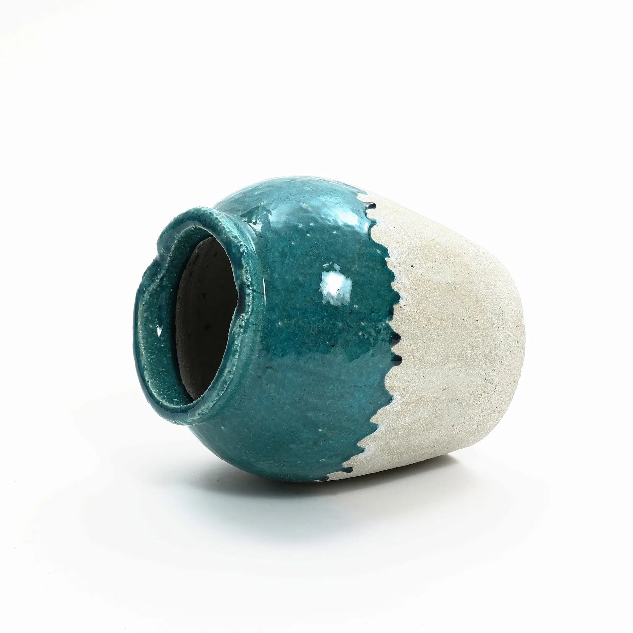 A ceramic vase with a glossy teal upper half and a textured beige lower half, embodying a rustic design, lies on its side against a white background.