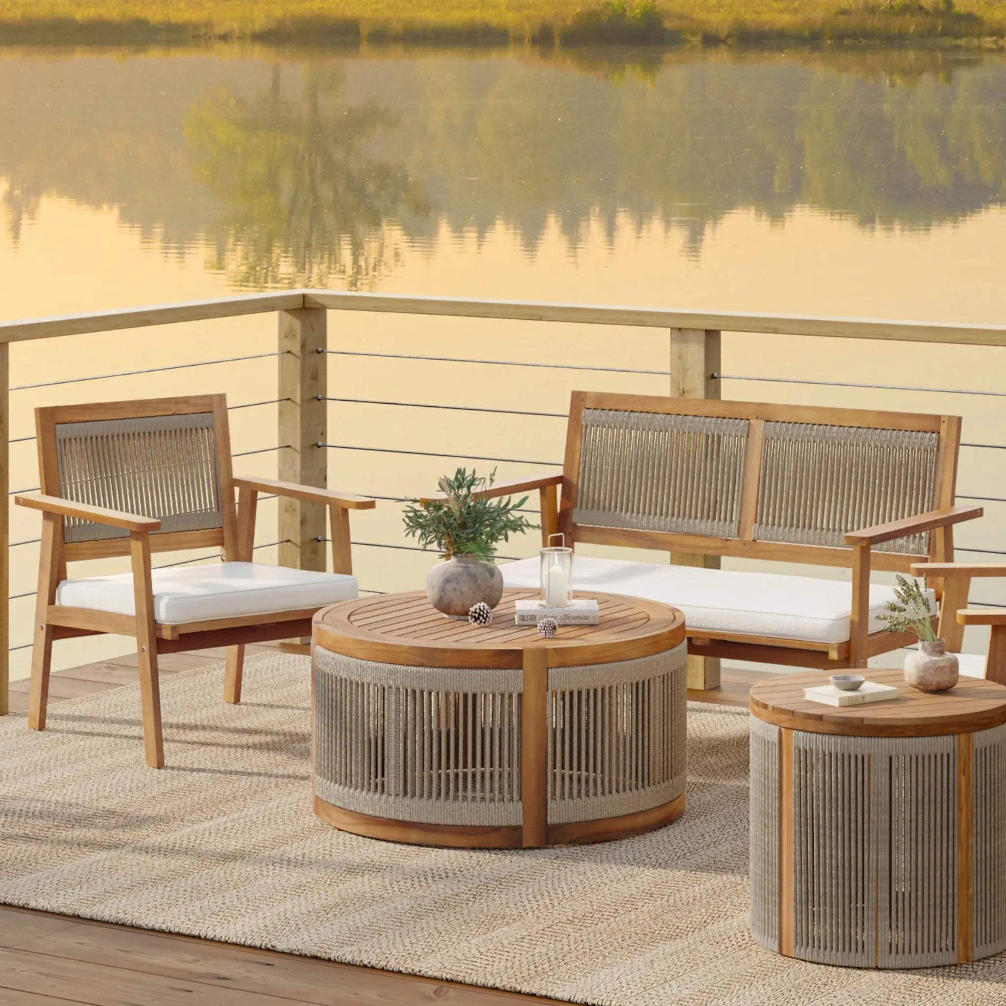 Outdoor patio set on a deck overlooking a calm lake, featuring an acacia wood sofa and chairs with plush cushions. The hand-woven rope circular coffee table and side table add charm to the decor, complemented by potted plants and candles. Perfect for indoor or outdoor relaxation.