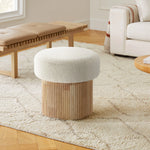 A cozy living room features the LuxenHome Mina boucle and wood storage ottoman with a textured white cushion and fluted base on a patterned rug. A light-colored sofa and wooden bench with a cushion in the background enhance the warm atmosphere.
