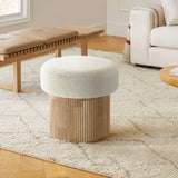 A cozy living room features the LuxenHome Mina boucle and wood storage ottoman with a textured white cushion and fluted base on a patterned rug. A light-colored sofa and wooden bench with a cushion in the background enhance the warm atmosphere.