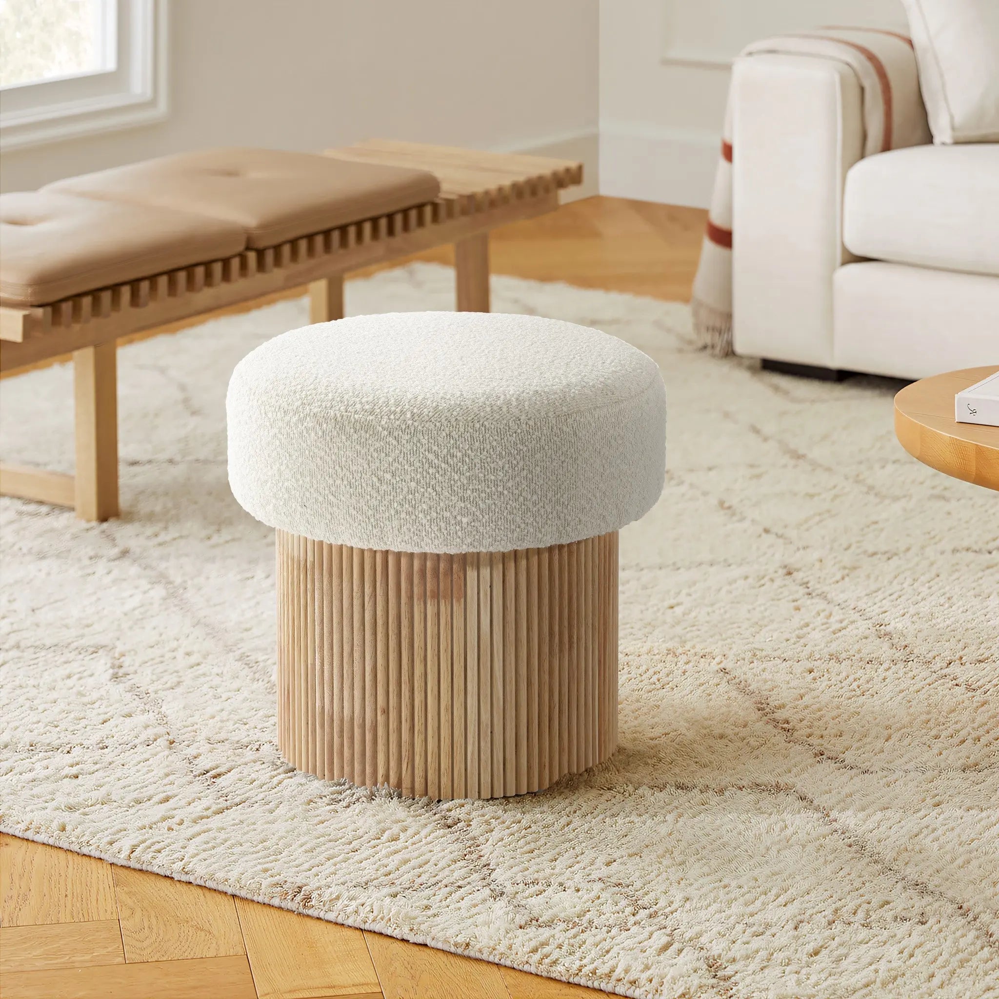 A cozy living room features the LuxenHome Mina boucle and wood storage ottoman with a textured white cushion and fluted base on a patterned rug. A light-colored sofa and wooden bench with a cushion in the background enhance the warm atmosphere.