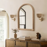 A decorative hanging mirror with an arched rectangular design features a wicker frame above the wooden console table. The table holds books, a vase with flowers, and sculptures. Two wall sconces are on either side of the mirror.