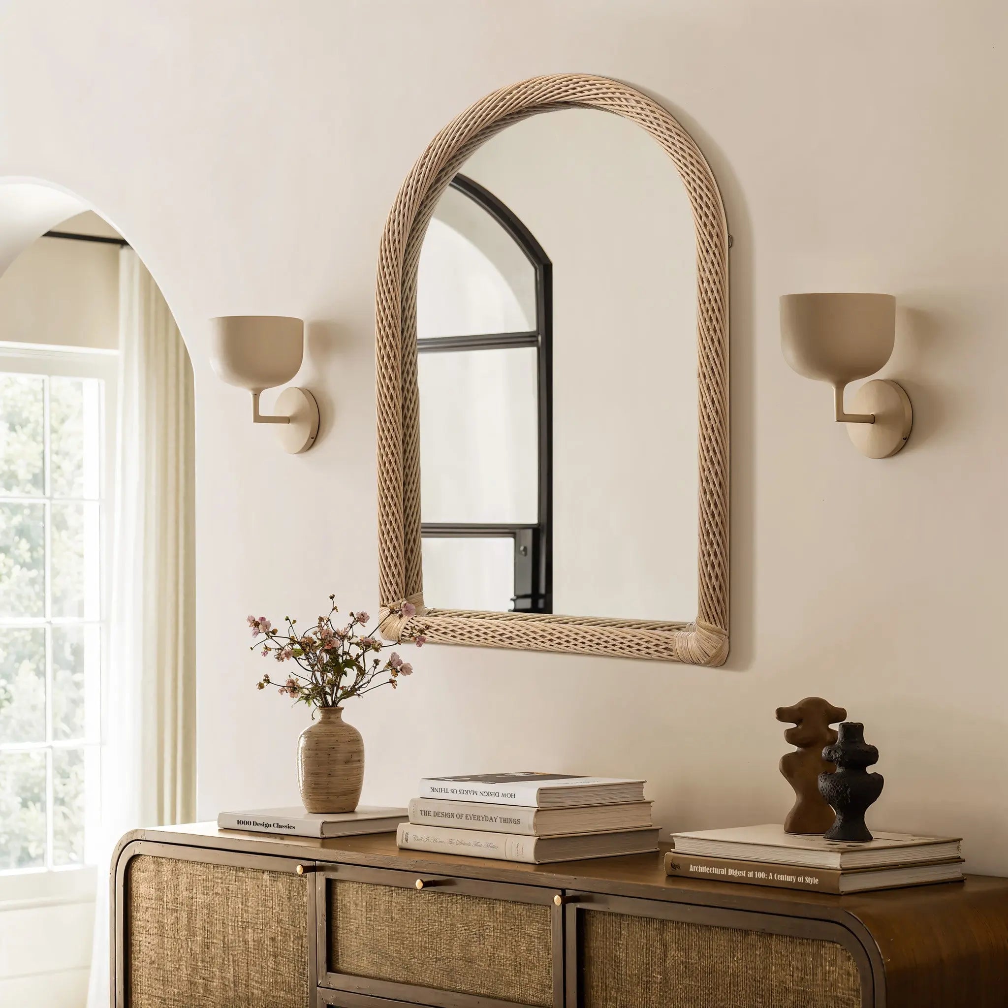 A decorative hanging mirror with an arched rectangular design features a wicker frame above the wooden console table. The table holds books, a vase with flowers, and sculptures. Two wall sconces are on either side of the mirror.