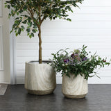 A stunning planter set featuring two large ceramic planters with an elegant ribbed texture, one showcasing a tall leafy tree and the other a mixed bouquet of purple and white flowers. They rest gracefully on a gray tiled floor against a crisp white paneled wall.