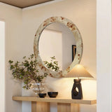 A round marble-framed mirror hangs above a wooden console, featuring a sleek modern lamp and a glass vase with greenery. The soft tones of the burlywood MDF frame complement the light wall, creating an elegant and harmonious setting.