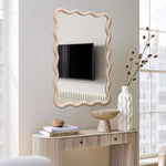 A wavy-framed decorative wall mirror reflects a wall-mounted TV and radiator. Below, a console table with a natural wooden frame holds a vase of dried flowers, a bowl, and a stack of books titled Best Furniture.