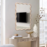 A wavy-framed decorative wall mirror reflects a wall-mounted TV and radiator. Below, a console table with a natural wooden frame holds a vase of dried flowers, a bowl, and a stack of books titled Best Furniture.