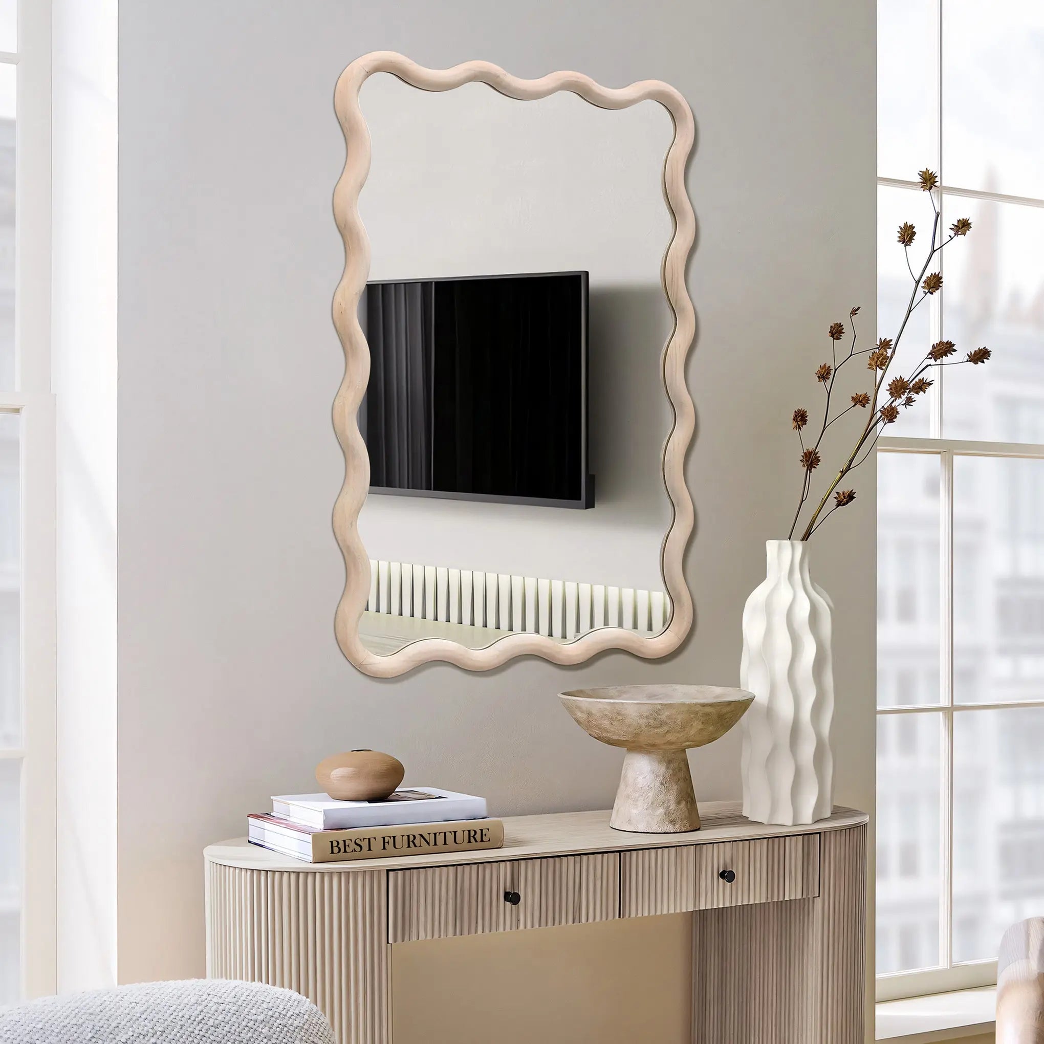 A wavy-framed decorative wall mirror reflects a wall-mounted TV and radiator. Below, a console table with a natural wooden frame holds a vase of dried flowers, a bowl, and a stack of books titled Best Furniture.