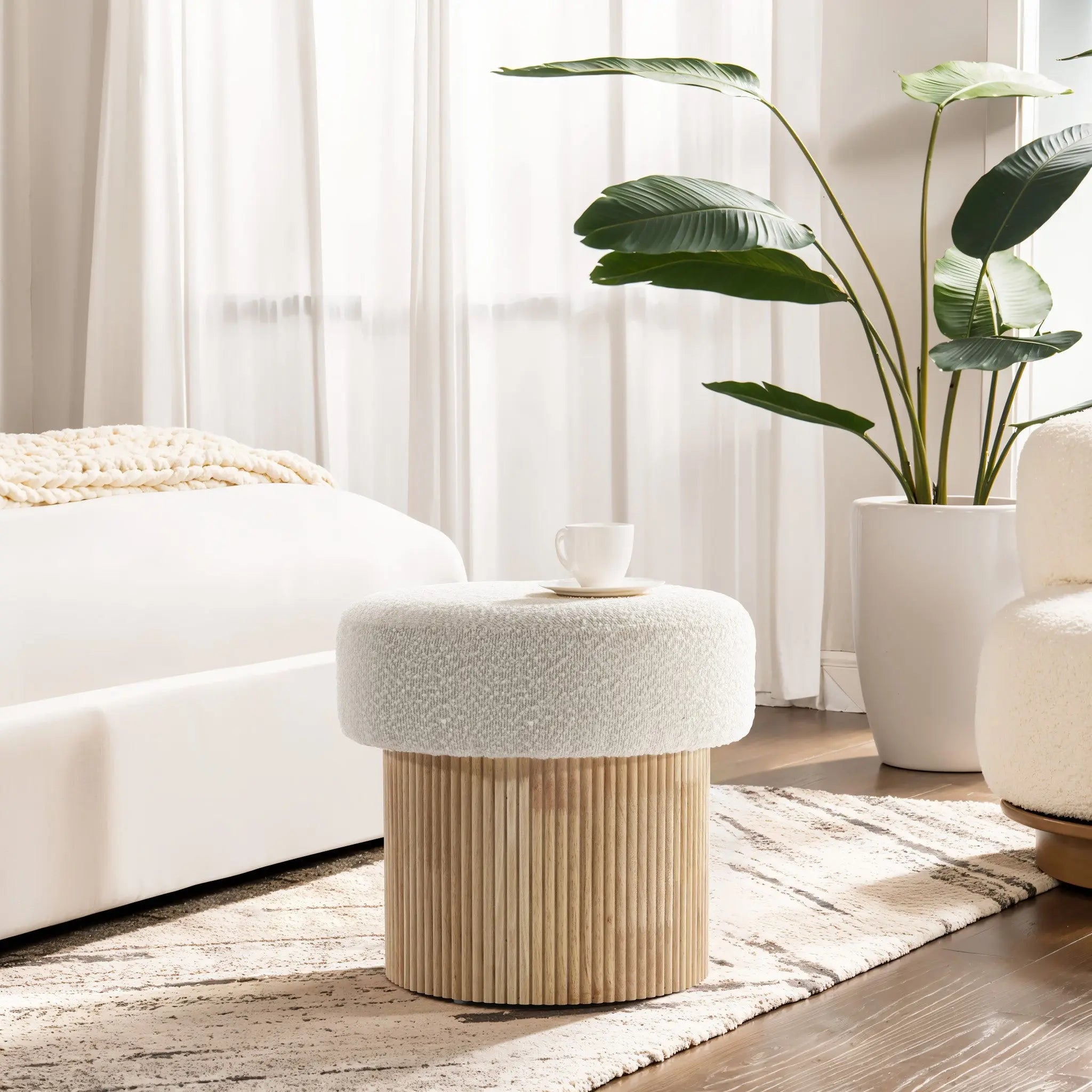 A cozy living room showcases the Mina boucle and wood storage ottoman by LuxenHome. It exudes elegance, topped with a white cup, sitting on a patterned rug next to a bed or sofa. In the background, white curtains frame a tall, leafy green plant.