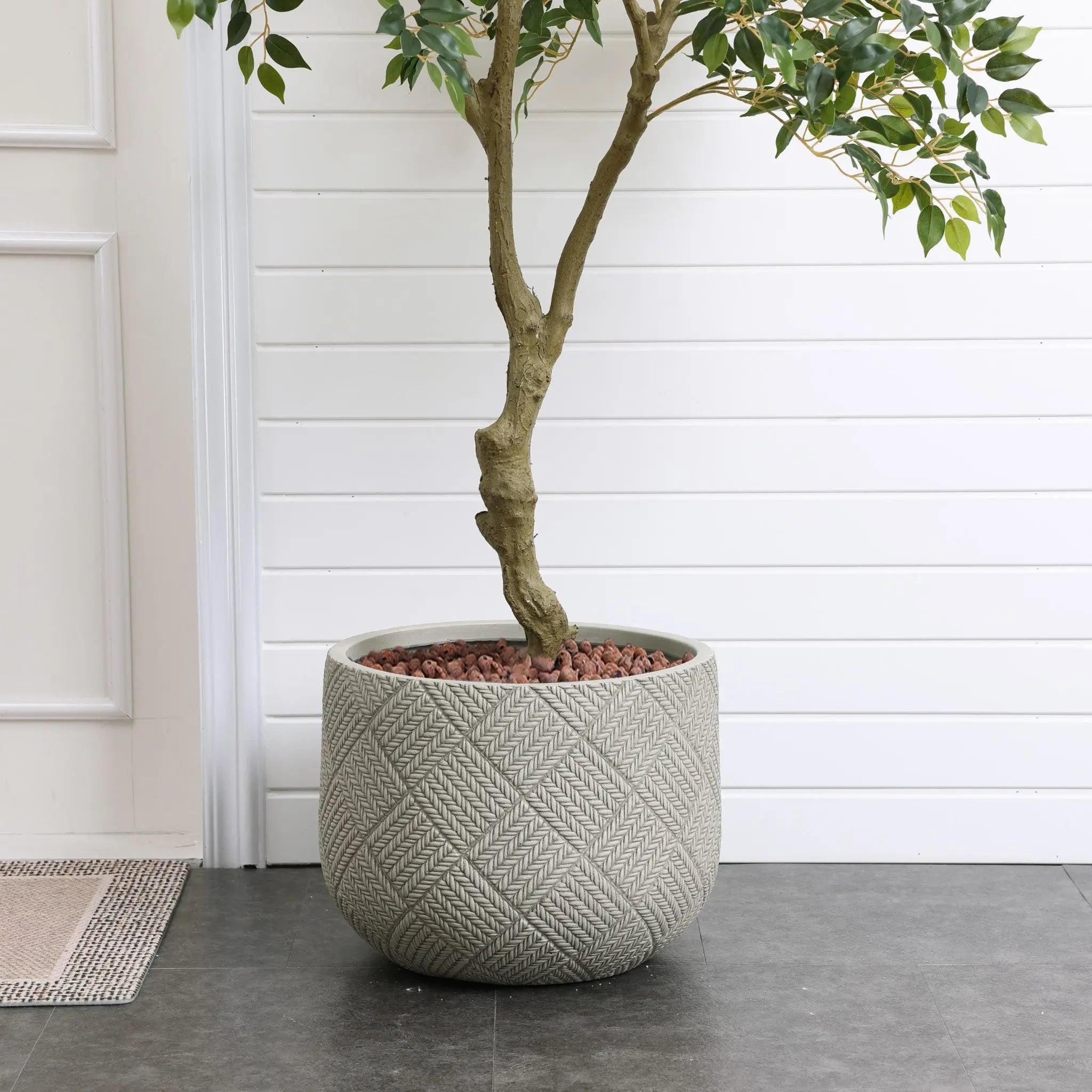 A tree with a thick trunk is planted in a large, textured MgO material planter filled with small red rocks, placed against a white paneled wall and a tiled floor.