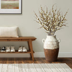 A minimalist entryway exudes rustic elegance with a wooden bench, a textured cushion, white sneakers neatly placed underneath, and LuxenHomes cream and brown terracotta urn vase with handles (18.5 h) on the right, holding white flowering branches beneath a framed picture on the light wall.