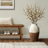 A minimalist entryway exudes rustic elegance with a wooden bench, a textured cushion, white sneakers neatly placed underneath, and LuxenHomes cream and brown terracotta urn vase with handles (18.5 h) on the right, holding white flowering branches beneath a framed picture on the light wall.