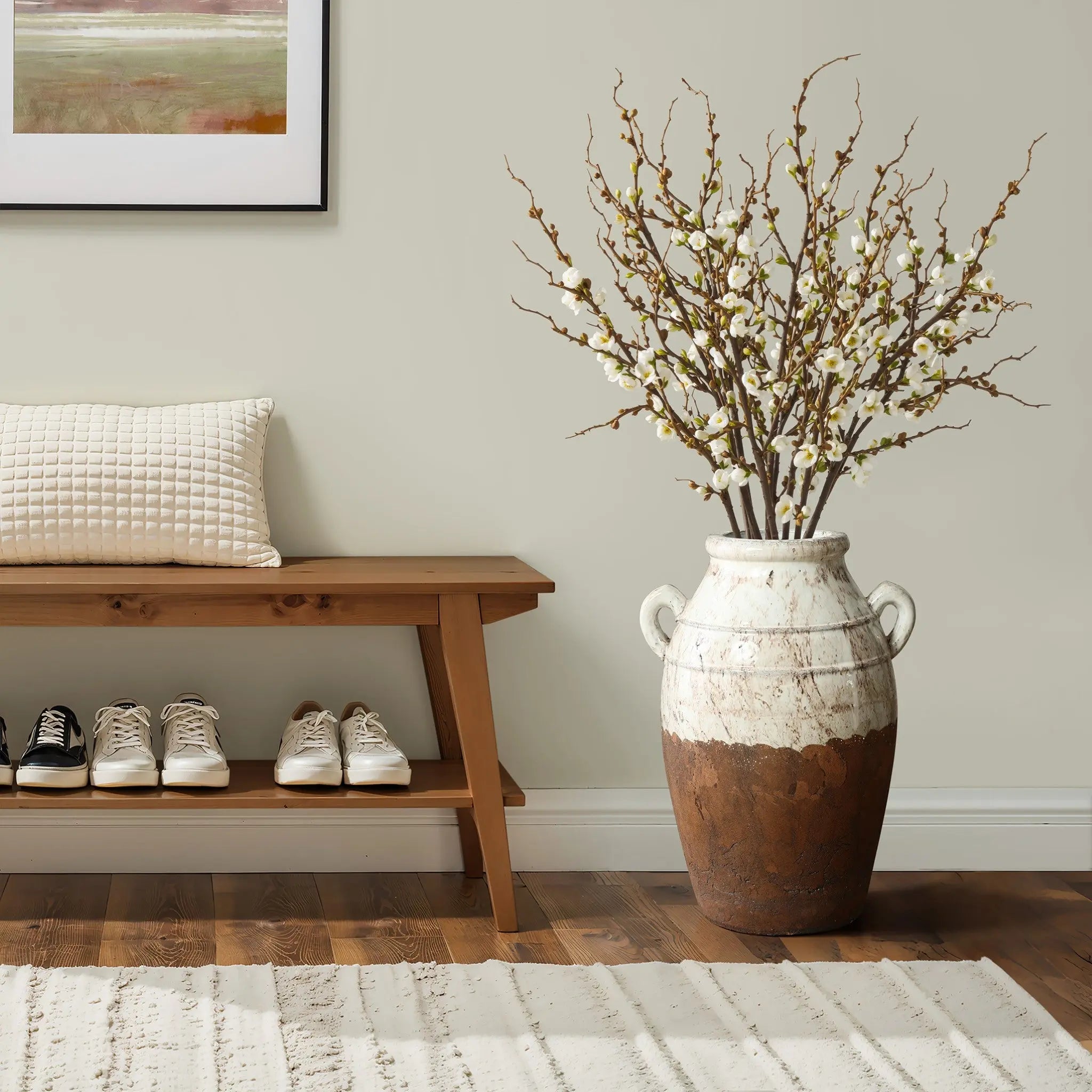A minimalist entryway exudes rustic elegance with a wooden bench, a textured cushion, white sneakers neatly placed underneath, and LuxenHomes cream and brown terracotta urn vase with handles (18.5 h) on the right, holding white flowering branches beneath a framed picture on the light wall.