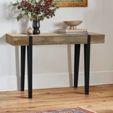 A modern farmhouse console table with an oak finish and black metal legs holds a vase with leafy branches, a bowl, and two stacked books, set against a white wall on a wooden floor.