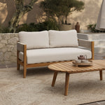 A Rope Sides Loveseat with white cushions sits beside an Outdoor Acacia Wood coffee table set topped with a teapot and cups, all arranged on a rug in a beautifully landscaped patio setting.