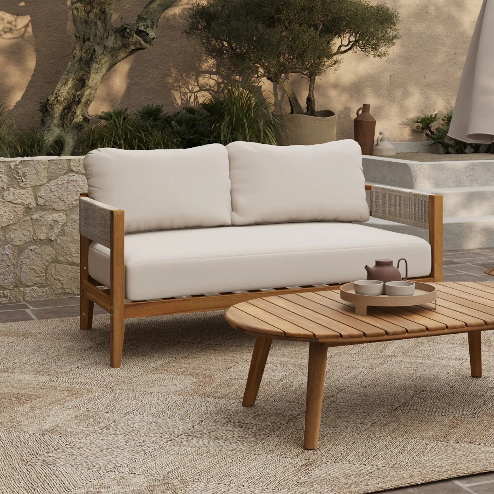 A Rope Sides Loveseat with white cushions sits beside an Outdoor Acacia Wood coffee table set topped with a teapot and cups, all arranged on a rug in a beautifully landscaped patio setting.