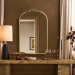 A cozy dining setup features a wooden table with wine glasses, black-candled candlesticks, grapes, and snacks. The Rêverie vintage gold accent metal frame arch mirror by LuxenHome enhances the wall decor as elegant curtains frame the arched window silhouette in the rooms corner.