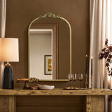 A cozy dining setup features a wooden table with wine glasses, black-candled candlesticks, grapes, and snacks. The Rêverie vintage gold accent metal frame arch mirror by LuxenHome enhances the wall decor as elegant curtains frame the arched window silhouette in the rooms corner.