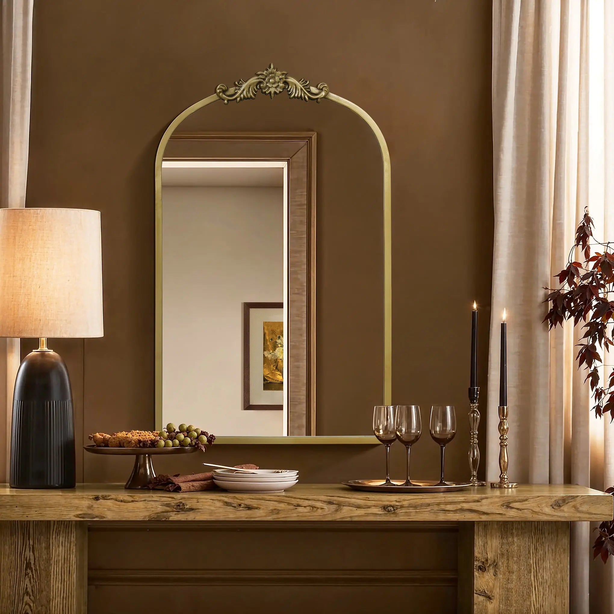 A cozy dining setup features a wooden table with wine glasses, black-candled candlesticks, grapes, and snacks. The Rêverie vintage gold accent metal frame arch mirror by LuxenHome enhances the wall decor as elegant curtains frame the arched window silhouette in the rooms corner.