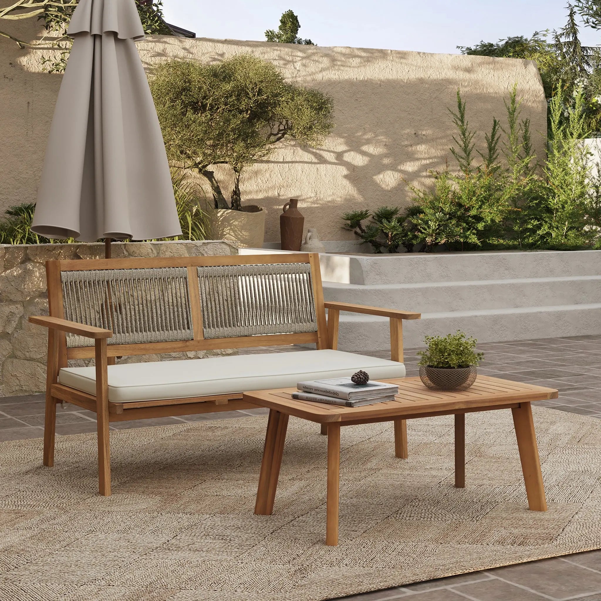 This modern minimalist furniture set features an FSC-certified Acacia wood bench with cushions and a table, all resting on a beige rug. Positioned against a stone wall, it’s complemented by lush plants and an umbrella, offering a serene outdoor retreat.