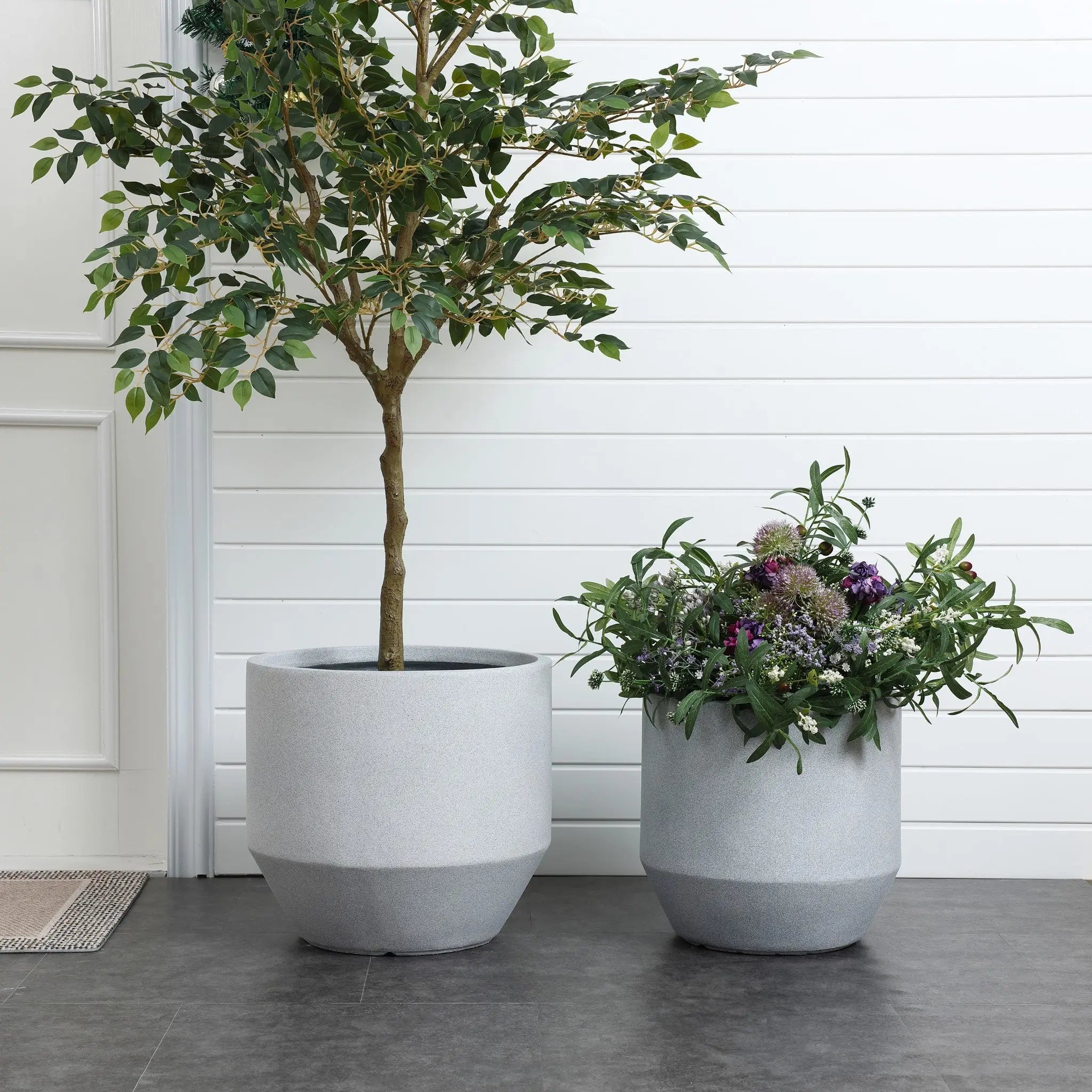 Two large, modern minimalist planters grace the space; the left one cradles a small tree, while the right overflows with purple and white blooms. Perfect for indoor and outdoor decor, these pots stand elegantly on a dark floor against a crisp white wall.