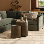 Living room with a green sectional sofa, two teardrop accent tables, decorative items, and books on the tables. A patterned pillow and a woven rug complete the rustic charm of this cozy setting.