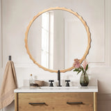 A round mirror with a scalloped rubberwood frame is above a bathroom sink. The vanity includes a wooden drawer, with the countertop adorned by a vase of flowers, a towel, and a soap dispenser.