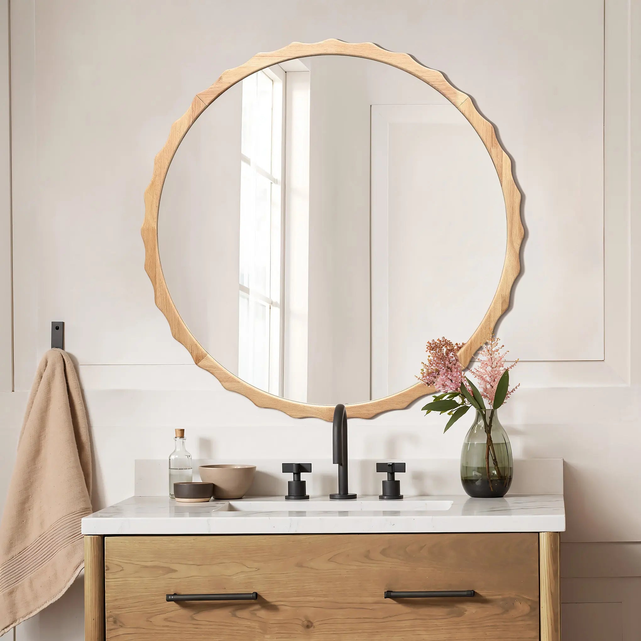 A round mirror with a scalloped rubberwood frame is above a bathroom sink. The vanity includes a wooden drawer, with the countertop adorned by a vase of flowers, a towel, and a soap dispenser.