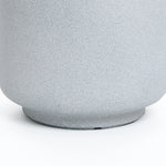 The image displays the lower part of a textured, light gray cylindrical object, possibly a weather-resistant planter or pot with a slightly wider base, resting on a smooth white surface.
