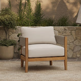 A cushioned Outdoor Acacia Wood Armchair with light beige upholstery sits on a textured outdoor rug, framed by a stone wall, potted plant, and lush greenery in the background.