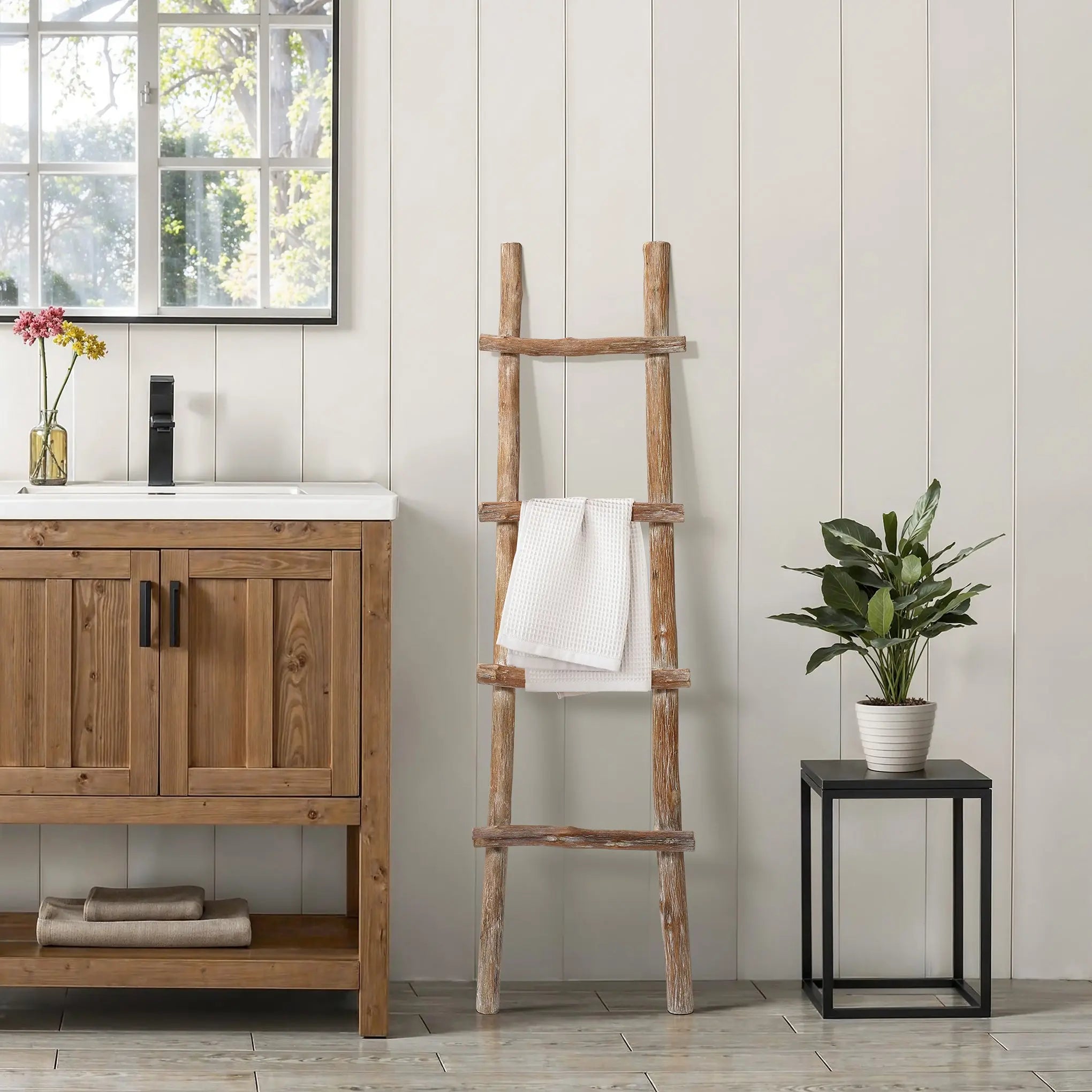 A solid wood blanket ladder leans against a white paneled wall, exuding rustic charm with a folded towel draped over it. To the right, modern functionality is showcased by a potted plant on a sleek black table next to a wooden sink cabinet.