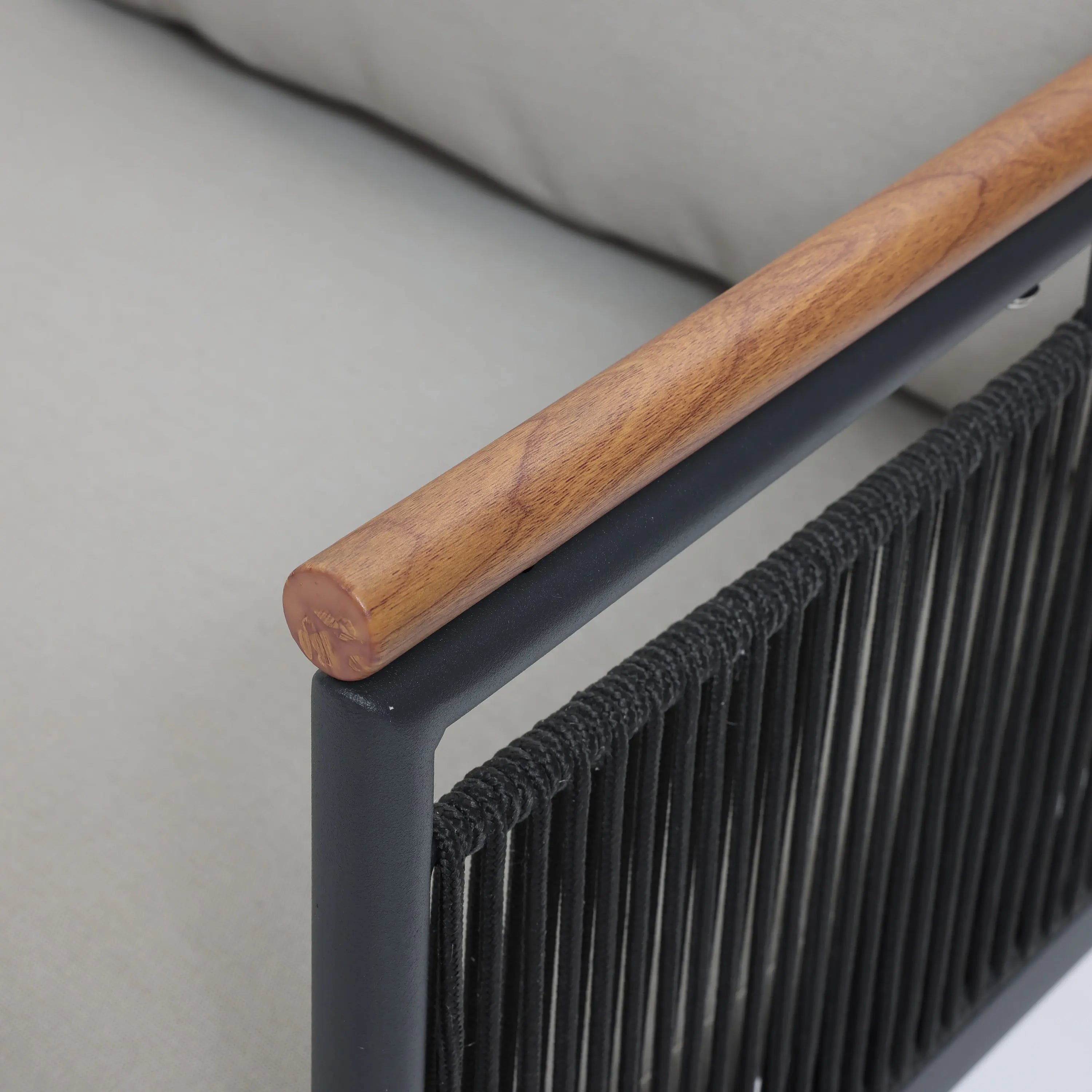 Close-up of a chair armrest featuring a wooden top, black metal frame, and woven black cord sides designed with subtle rope vertical stripes. In the background, a light gray cushion complements the outdoor seating set beautifully.