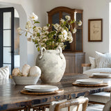 A rustic dining room showcases a wooden table set with white plates and woven chargers. At the center is the LuxenHomes rustic gray and white terracotta vase with handles, adding elegance. Large white decor objects, a wooden cabinet, and neutral decor complete the look.