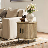 A cozy living room corner features a beige sofa adorned with patterned cushions. LuxenHomes Aria oak finish nightstand with a slatted sliding door holds a vase of white roses and decorative bowls, all set on a patterned rug. Natural light streams in from nearby sliding doors.