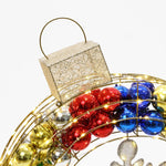 A festive Christmas wreath with red, blue, and gold ornaments is topped by a glittery rectangular decoration, adorned with warm white outdoor lights.