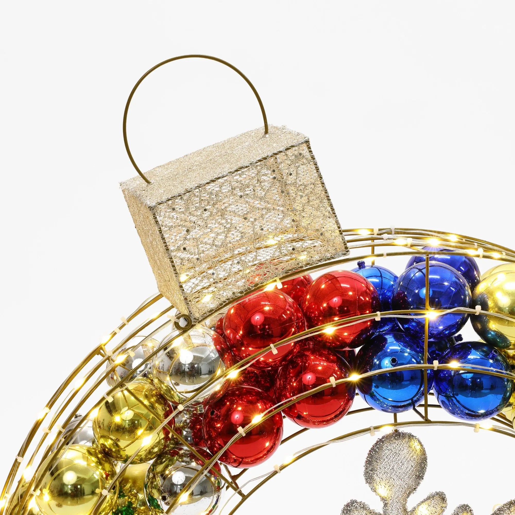 A festive Christmas wreath with red, blue, and gold ornaments is topped by a glittery rectangular decoration, adorned with warm white outdoor lights.