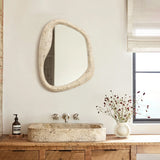 The bathroom features a rustic stone basin paired with an oval wall mirror in an abstract style. A wooden counter complements the black soap dispenser, alongside a vase of red and white flowers. White blinds shade the window, enhancing this elegant home decor oasis.