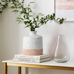A bottle-like design stoneware vase with green branches sits on a stack of books, complementing the clear glass vase on the table.
