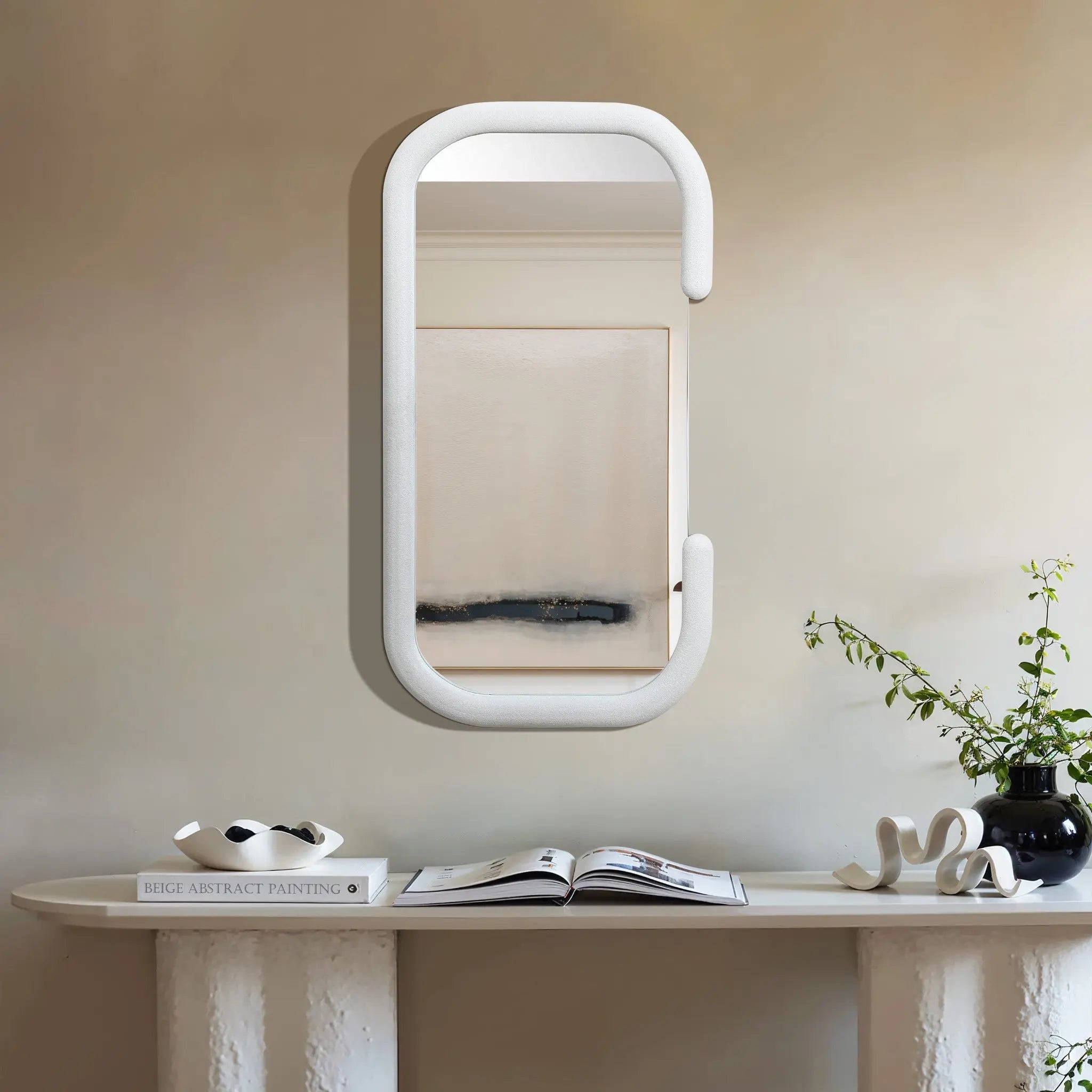 A touch of modernist elegance defines this space, featuring a decorative wall mirror on a beige expanse. Its rectangular surface reflects abstract art, while below, a pristine white table holds an open book, tasteful decor items, and a vase brimming with lush greenery.