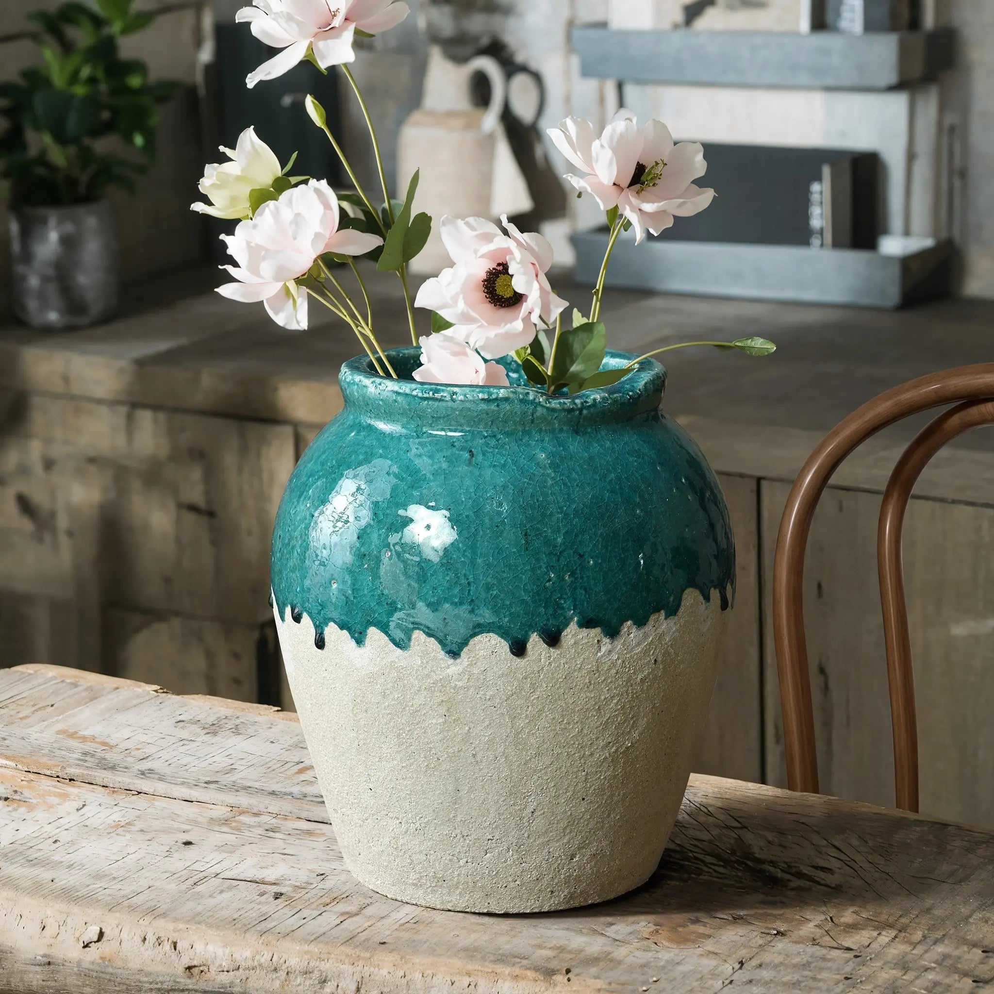 A rustic design comes to life with a teal and beige ceramic vase holding pink flowers on a wooden table. A chair and shelves add charm in the background, accentuating the cozy setting.