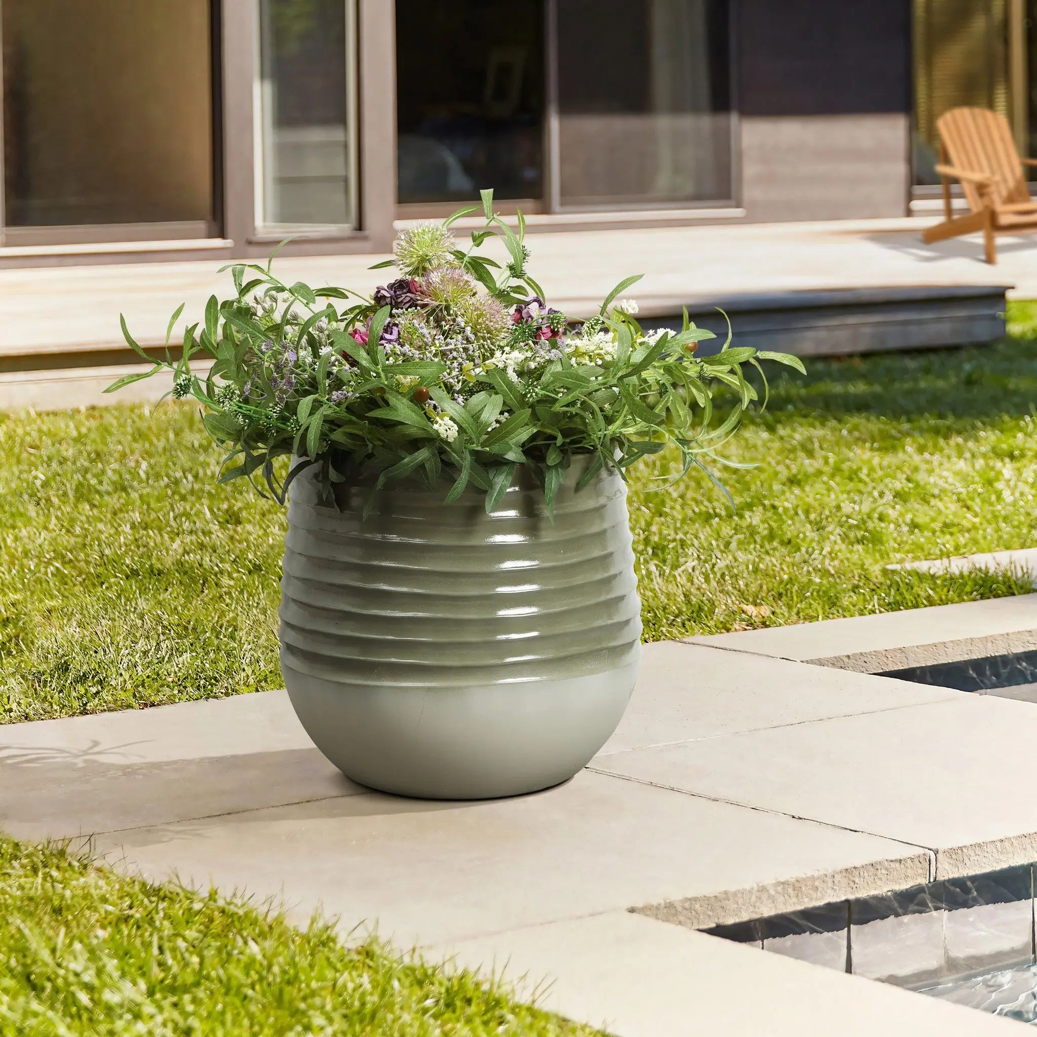 A round planter with a handy drainage hole, showcasing green and purple flowers, graces a stone patio beside a lawn. A wooden chair sits in the background, seamlessly blending indoor and outdoor environments.