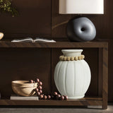 A wooden shelf showcases a modern round vase adorned with beads, alongside a bowl of round objects, a black abstract sculpture, a closed book, and a minimalist lamp. The arrangement embodies minimalist design while adding elegance to the space.